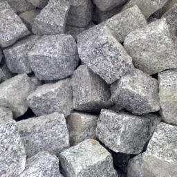 Stos szarych, surowych kostek granitowych o nieregularnych kształtach i widocznej fakturze, przygotowanych do ułożenia nawierzchni.